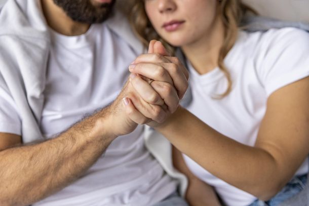 Imagen en primer plano de una pareja joven, enfocada en sus manos entrelazadas, que transmiten conexión y apoyo mutuo. Ambos visten camisetas blancas y están sentados muy próximos, con una actitud de cercanía emocional. Aunque sus rostros aparecen parcialmente desenfocados, se percibe en sus expresiones una actitud reflexiva e íntima. Esta escena representa simbólicamente el trabajo emocional en pareja, las dificultades relacionales y el acompañamiento psicológico en contextos de terapia de pareja, uno de los servicios ofrecidos por Raquel Egido, psicóloga profesional.