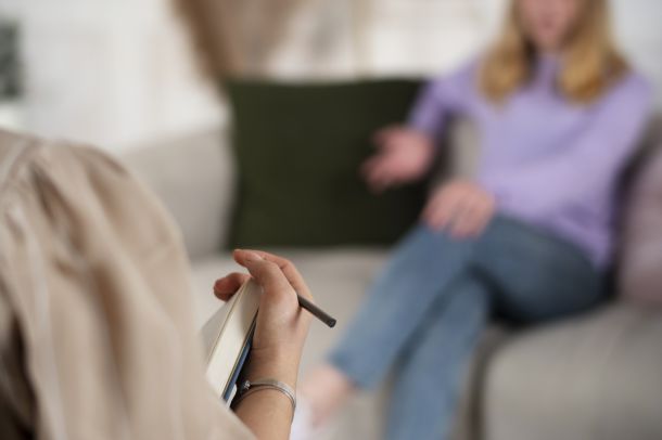 Fotografía desenfocada de una paciente sentada en un sofá durante una sesión de terapia, frente a Raquel Egido, psicóloga profesional, cuya mano en primer plano sostiene un cuaderno y un bolígrafo. La paciente, con suéter lila y pantalones vaqueros, gesticula mientras conversa. El fondo muestra cojines oscuros y un entorno cálido y neutro, típico de una consulta terapéutica. La escena refleja el proceso de escucha activa, acompañamiento emocional y vínculo profesional que se desarrolla en una sesión de psicoterapia.