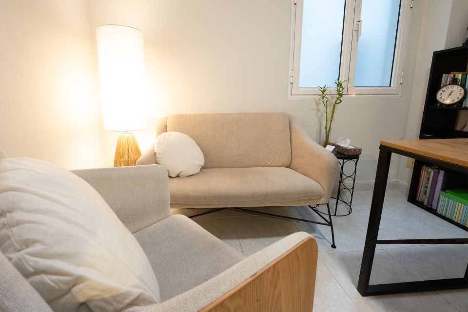 Vista parcial de la consulta de Raquel Egido, psicóloga profesional, centrada en la zona de espera o atención al paciente. En primer plano se observa un sillón beige claro con cojín blanco, y al fondo, un sofá del mismo estilo con otro cojín redondo. A la izquierda, una lámpara de pie con pantalla blanca emite una luz cálida que genera una atmósfera acogedora y relajante. Junto al sofá hay una pequeña mesa auxiliar con pañuelos y una planta decorativa de bambú. La escena transmite calma, intimidad y comodidad, elementos esenciales en un espacio terapéutico.