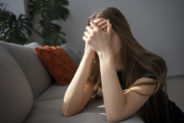 Imagen de una mujer sentada en un sofá gris, con un cojín naranja al fondo, en un entorno doméstico o terapéutico. Apoya ambos codos sobre sus piernas y mantiene las manos entrelazadas sobre el rostro, transmitiendo claramente una sensación de tensión, agotamiento emocional o ansiedad. Su cabello largo y su postura corporal reflejan un momento de bloqueo o sobrecarga mental. La escena representa simbólicamente el impacto del estrés en la vida diaria, un tema abordado en las sesiones de terapia con Raquel Egido, psicóloga profesional, quien ofrece herramientas específicas para identificar, reducir y afrontar el estrés y la ansiedad de forma terapéutica y efectiva.