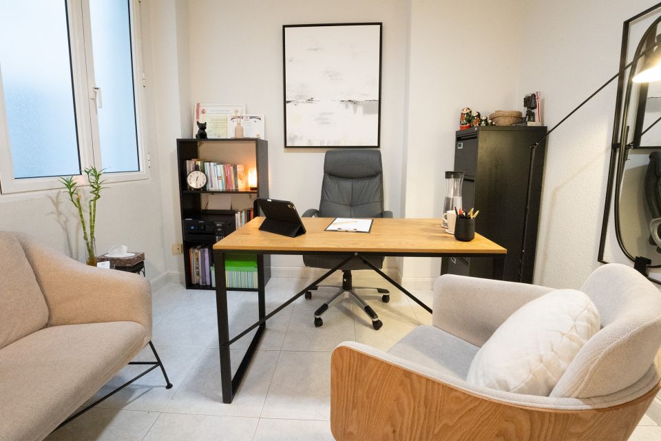 Imagen de la consulta psicológica de Raquel Egido, psicóloga profesional. La estancia muestra un ambiente acogedor y ordenado. En el centro hay un escritorio de madera con patas negras, sobre el cual descansan una carpeta, una tablet, un vaso con lápices y una jarra de agua con una taza. Detrás del escritorio se encuentra una silla ergonómica de oficina, y frente a ella, dos sillones confortables para pacientes. A la izquierda, una estantería con libros, diplomas y una pequeña lámpara crean un ambiente cálido y profesional. Una ventana con cristales traslúcidos permite la entrada de luz natural, mientras que una lámpara de pie aporta iluminación adicional. Todo el conjunto transmite calma, profesionalidad y bienestar.