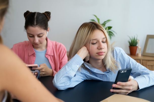 Imagen de dos adolescentes sentadas en una mesa, cada una centrada en su teléfono móvil. La joven en primer plano, de cabello rubio y blusa de rayas azules, apoya la cabeza en una mano con expresión seria o ausente. La otra chica, con camisa rosa y auriculares, también mira su dispositivo. Al fondo se observan plantas decorativas y mobiliario neutro. La escena transmite una sensación de desconexión emocional, aislamiento social o posible saturación digital, temas comunes en la adolescencia actual y habitualmente abordados en procesos de orientación psicológica.