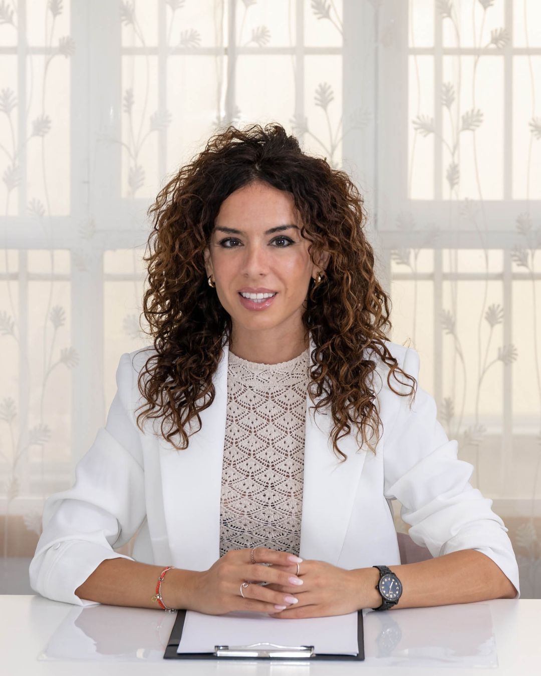 Raquel Egido, psicóloga profesional, aparece sentada frente a un escritorio blanco con un portafolios de documentos. Lleva el cabello rizado suelto, y viste una chaqueta blanca sobre una blusa de encaje claro. Sus manos están relajadas y entrelazadas sobre la mesa, mostrando una actitud tranquila y profesional. Al fondo, se aprecia una ventana con cortinas blancas translúcidas decoradas con un sutil estampado vegetal que deja pasar luz natural suave. La imagen transmite cercanía, confianza y profesionalidad, representando un entorno seguro y acogedor para la consulta psicológica.