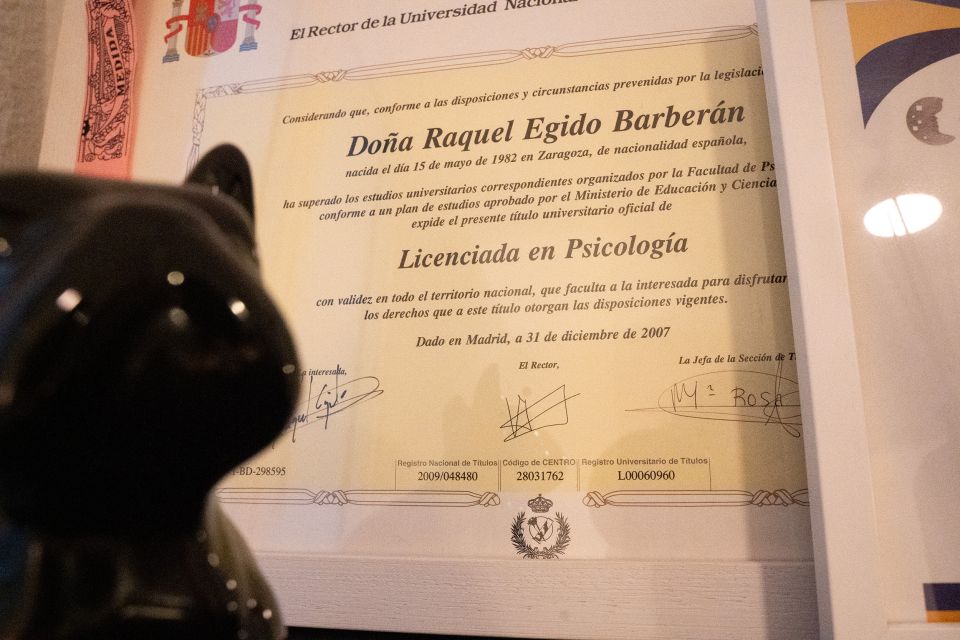 Fotografía en primer plano del título universitario oficial de Raquel Egido Barberán, emitido por la Universidad Nacional a Distancia (UNED). El diploma certifica que ha completado los estudios correspondientes para obtener el título de Licenciada en Psicología, con validez en todo el territorio nacional. La fecha de expedición es el 31 de diciembre de 2007 en Madrid. En la imagen también se aprecia parcialmente una figura decorativa de un gato negro brillante en primer plano, desenfocada, que da un toque personal y acogedor al espacio. El encuadre sugiere que el documento está enmarcado y expuesto en la consulta psicológica, reforzando la confianza y profesionalidad del entorno.